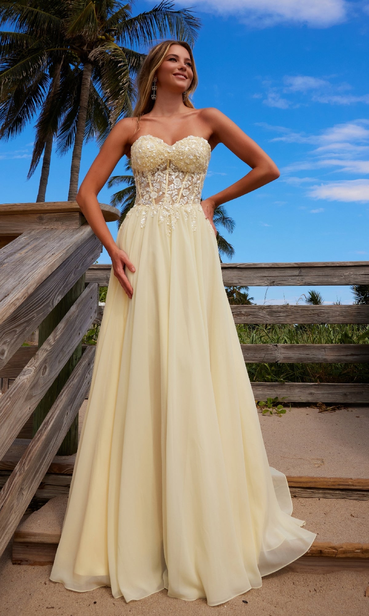 A woman in the Nox Anabel C1617 Strapless Sweetheart Prom Dress with a lace bodice and floor-length light yellow skirt stands on a wooden walkway at the beach, framed by palm trees and a blue sky.